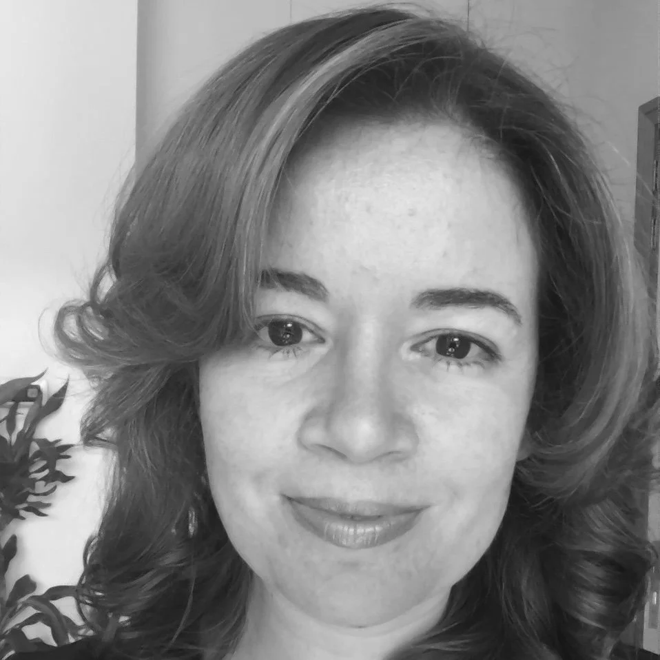 Black and white portrait of a woman with wavy hair smiling at the camera, with plants visible in the background.