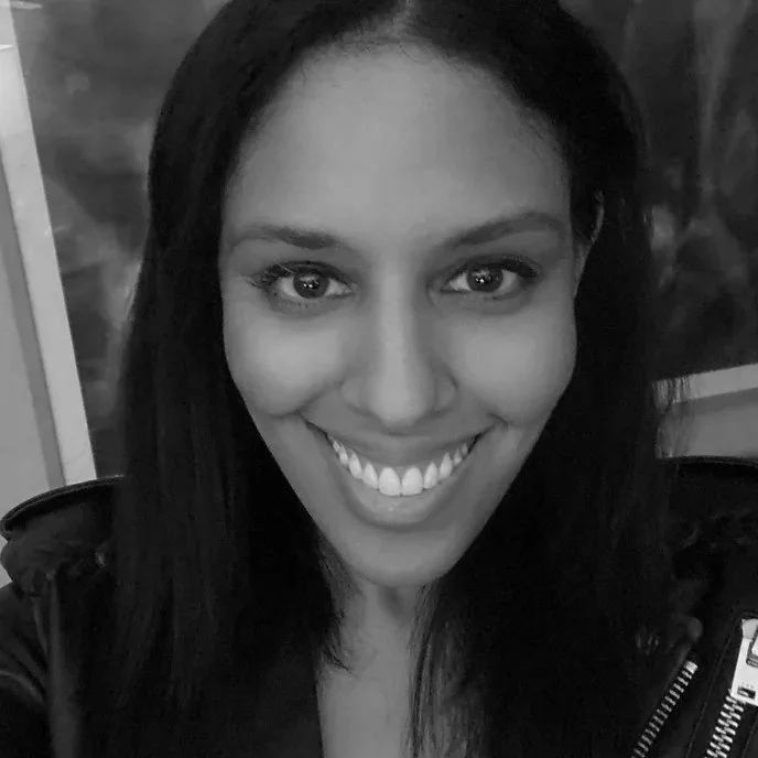 A woman smiling with white teeth, dark hair, and light eyes, wearing a dark jacket.