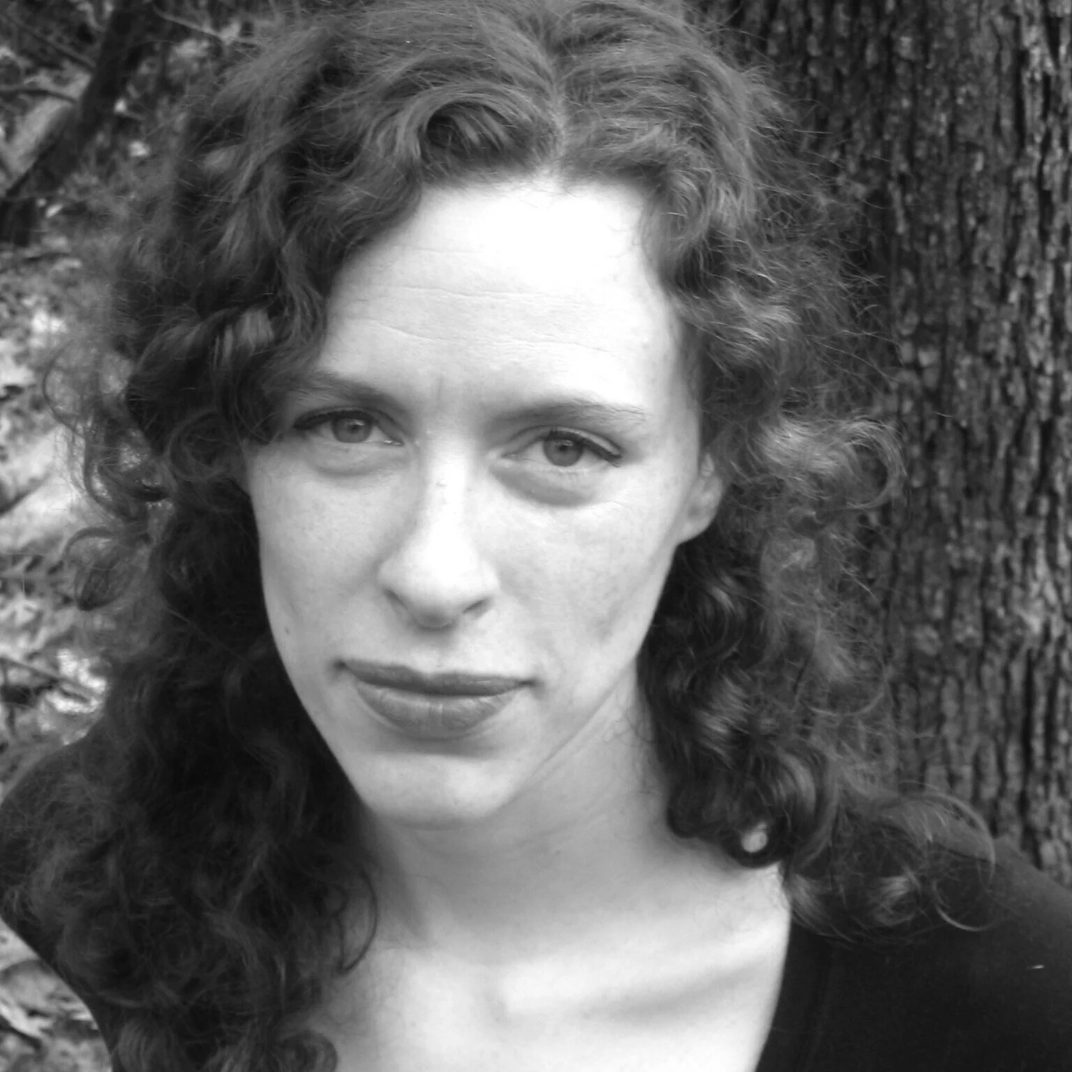 A black and white portrait of a woman with curly hair, standing outdoors in front of a tree.