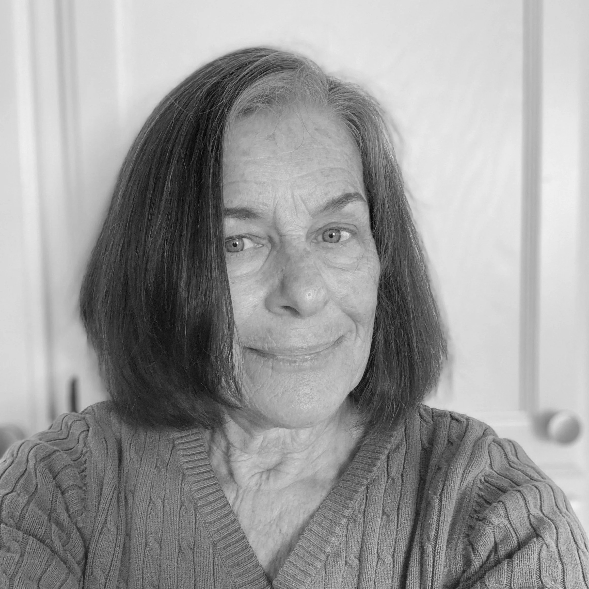 A black-and-white photograph of an older woman with shoulder-length dark hair, wearing a ribbed sweater, smiling gently, with a wooden cabinet in the background.