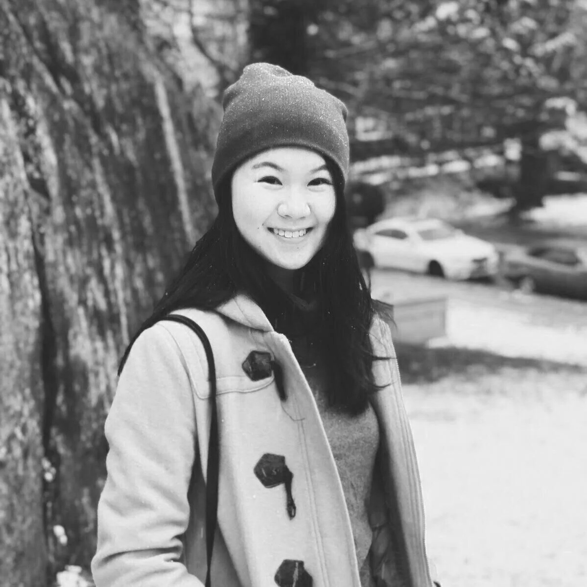 A smiling woman wearing a beanie and jacket outdoors near a tree with parked cars in the background.