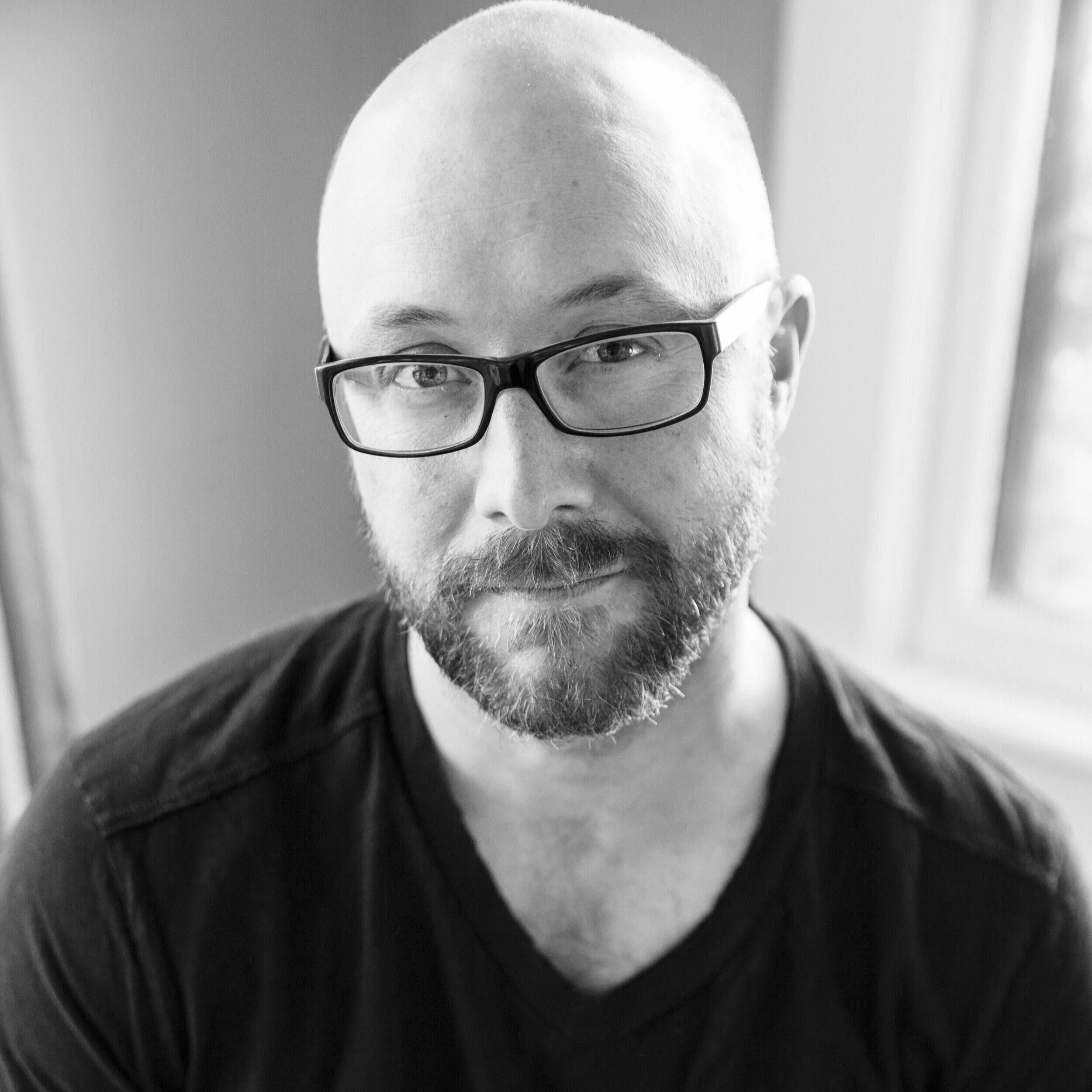 Black and white portrait of a bald man with a beard wearing glasses and a black shirt, looking at the camera near a window.