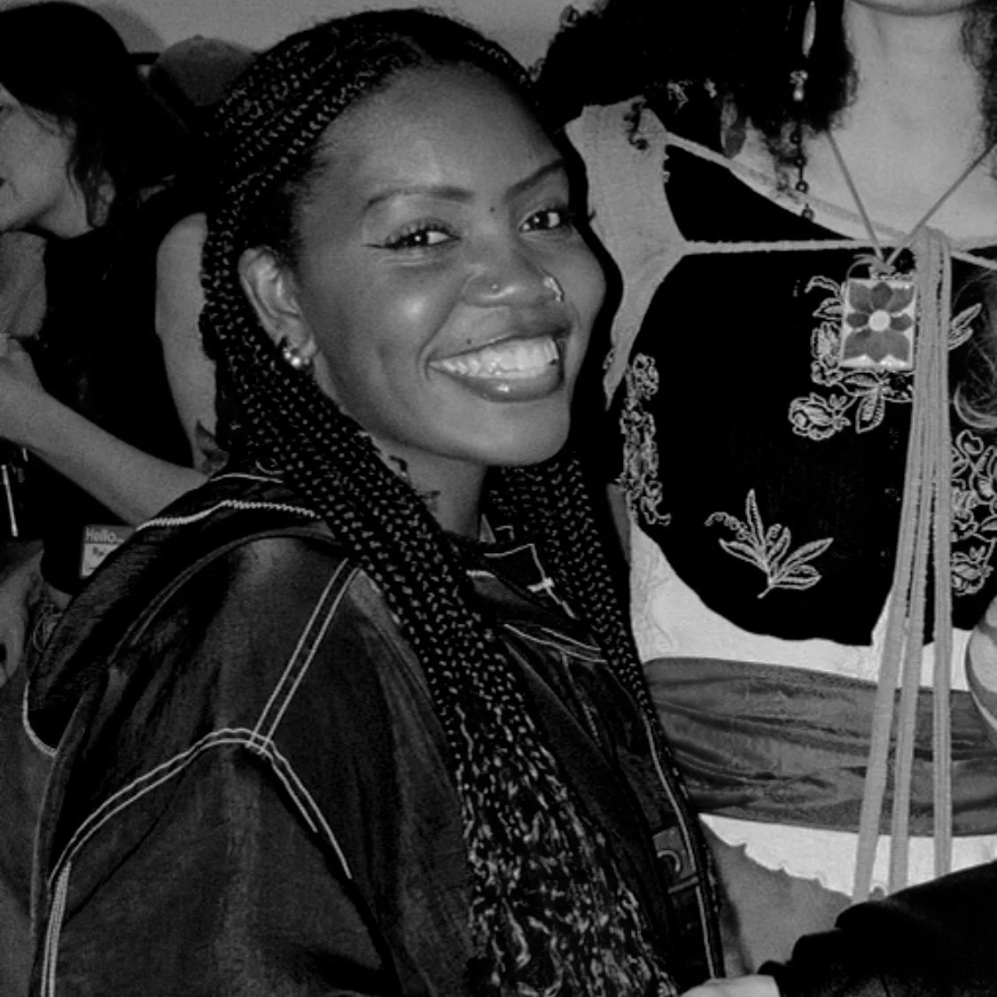 A smiling woman with braided hair, wearing earrings and a dark jacket with light stitching, posing at a social gathering.