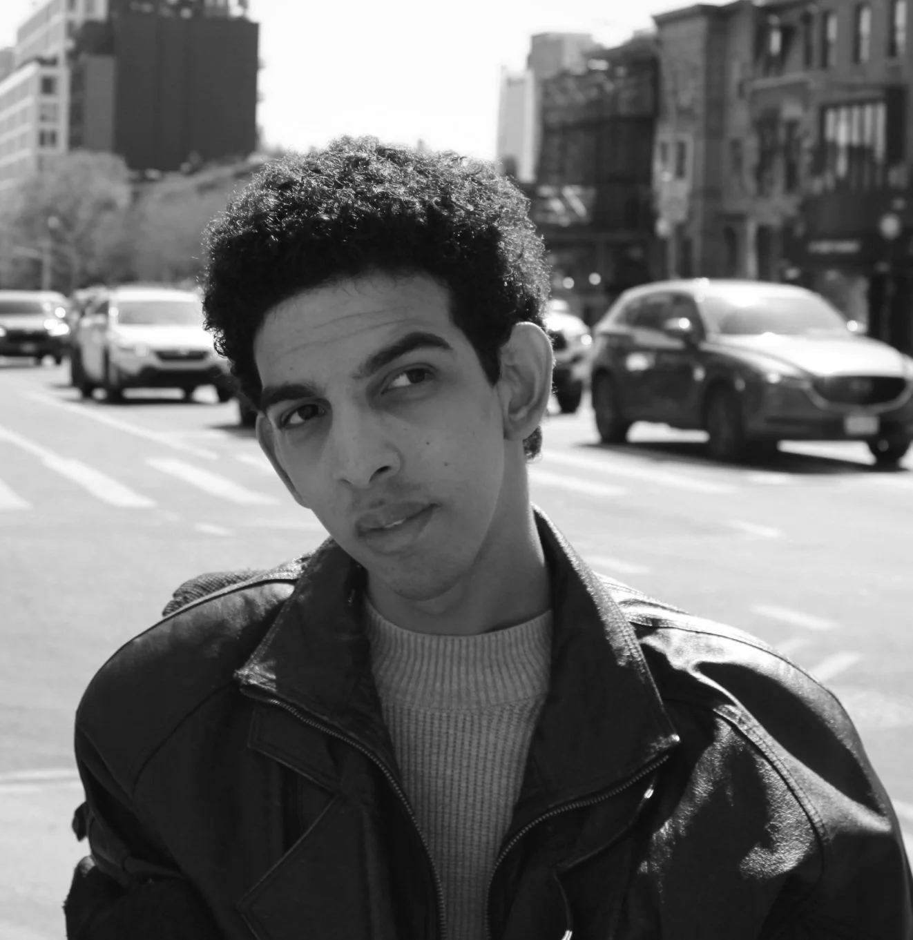 A young man with curly hair wearing a leather jacket and a sweater, standing on a city street with cars and buildings in the background.