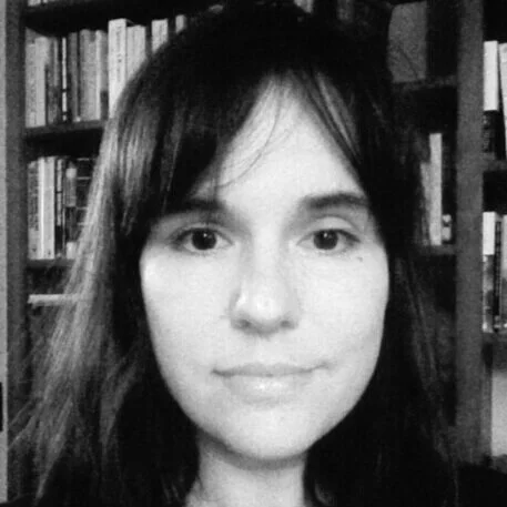 Black and white photo of a young woman with shoulder-length dark hair, standing in front of a bookshelf filled with books.