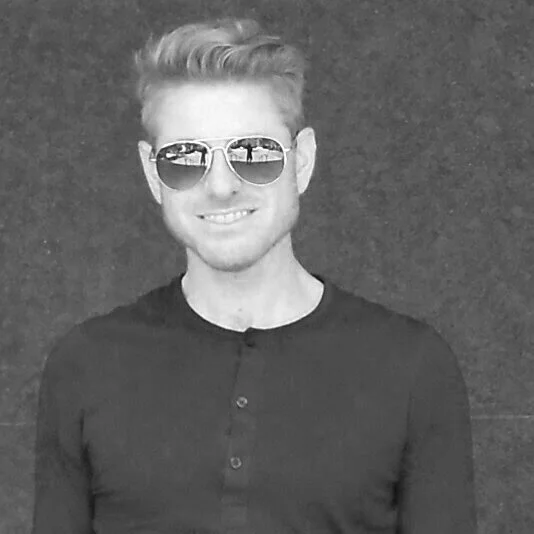 A young man with light-colored hair styled upward, wearing aviator sunglasses and a black long-sleeve shirt, smiling at the camera against a dark background.