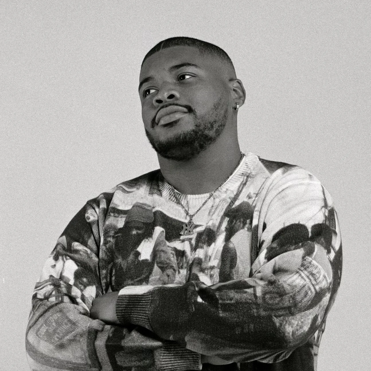 A black and white photo of a man with short hair, a beard, and an earring, crossing his arms and looking to the side.