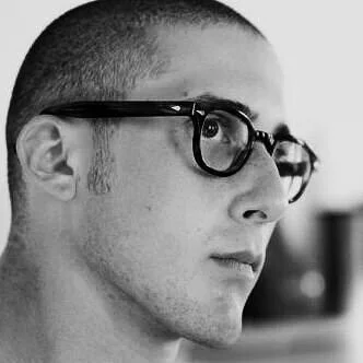 Black and white close-up of a young man with glasses, shaved head, and a thoughtful expression.