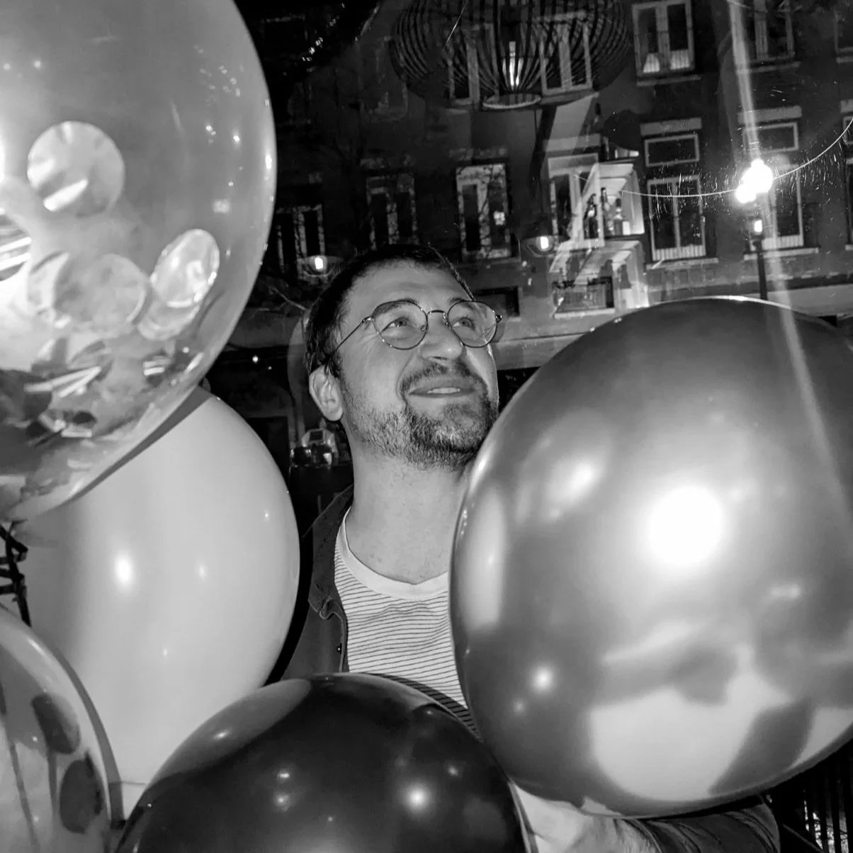A man with glasses and facial hair smiling while surrounded by balloons, with a city street visible through the window behind him at night.