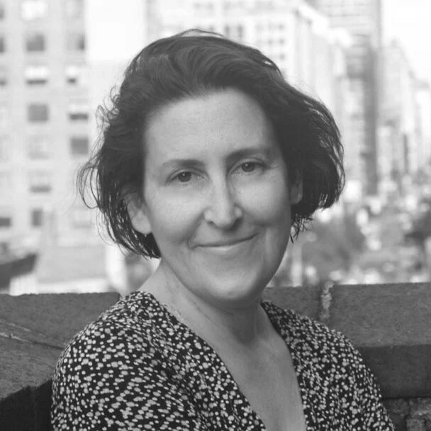 Black and white photo of a woman with short, wavy hair smiling outdoors, with city buildings in the background.