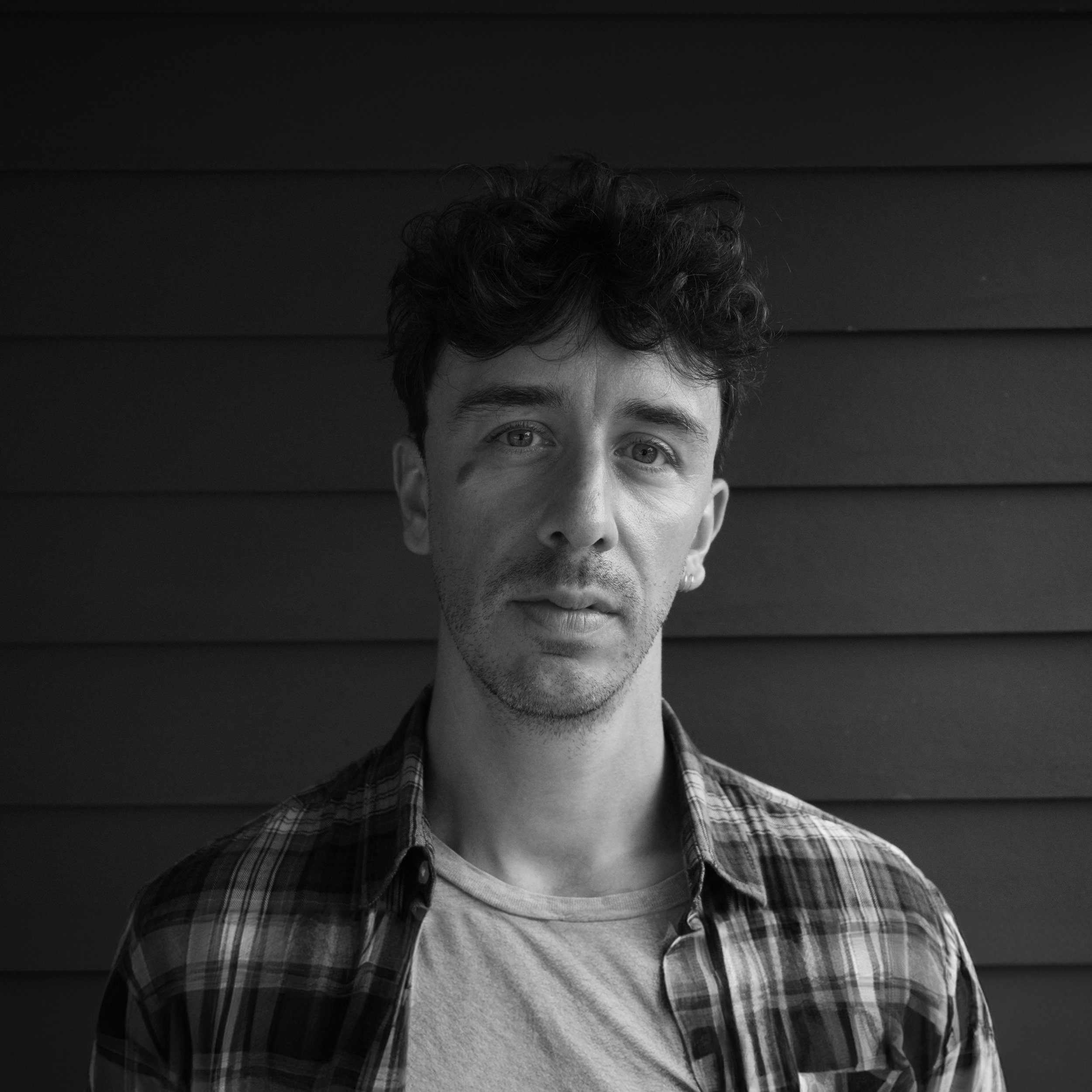 Black and white portrait of a young man with curly hair, tattoos, and earrings, wearing a plaid shirt and t-shirt, standing against a wooden wall.