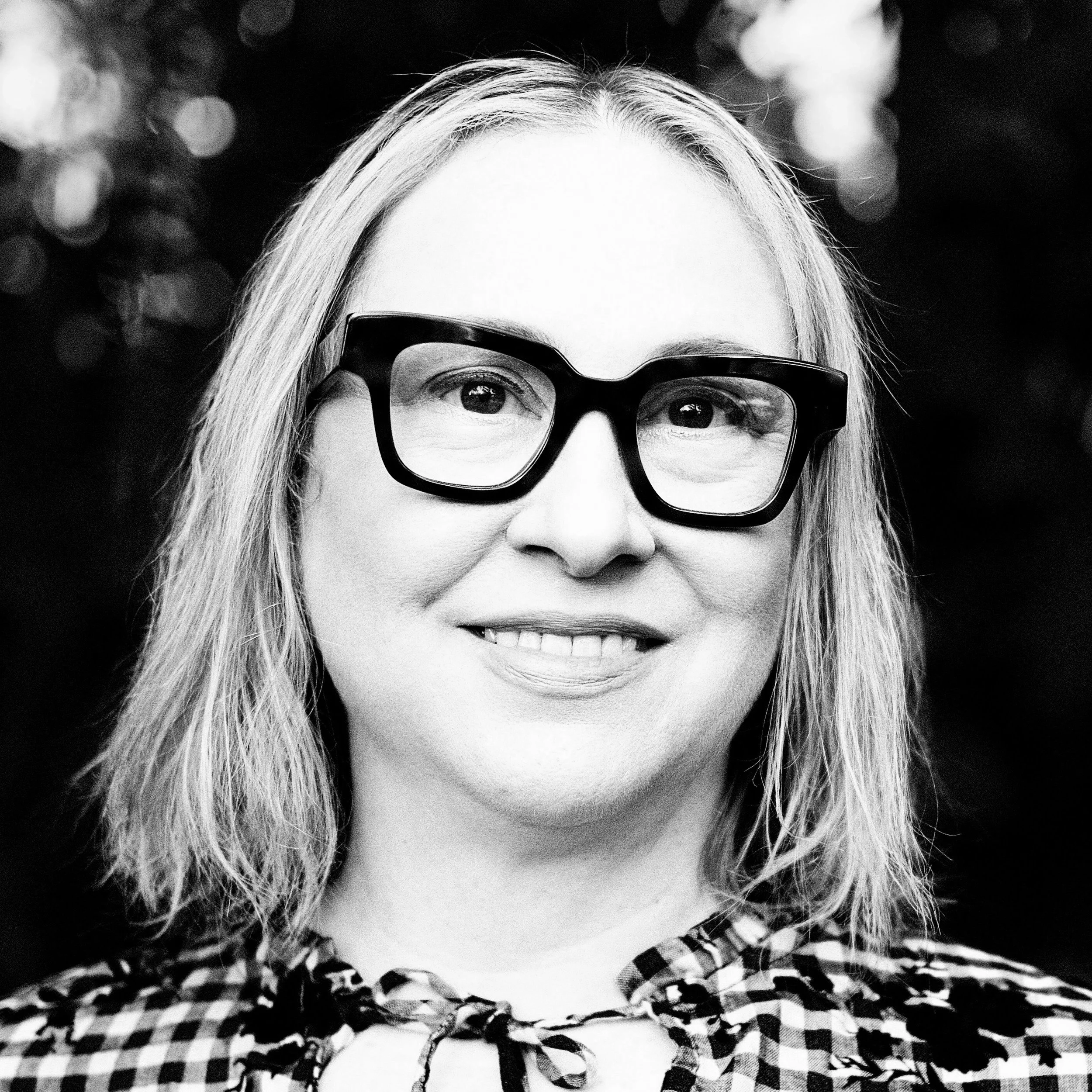 Black and white portrait of a woman wearing large glasses, smiling, with shoulder-length hair and a checkered top with a bow tie.