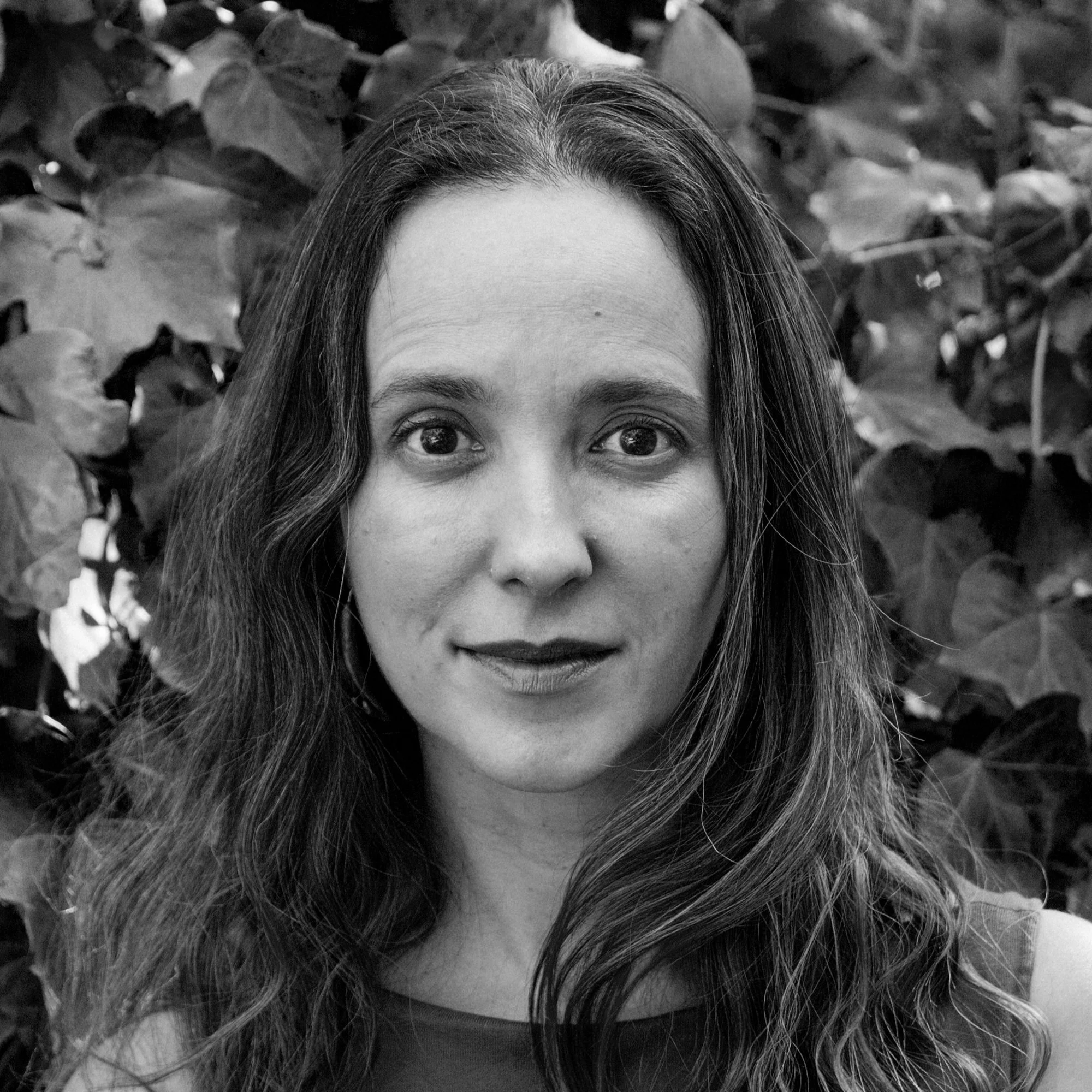 Black and white portrait of a woman with long wavy hair standing in front of leafy background.