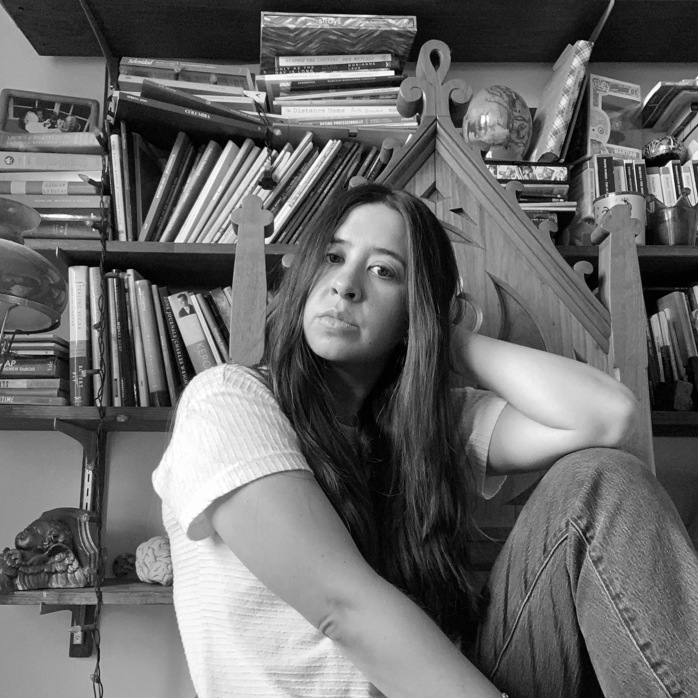A woman sitting in front of a large bookshelf filled with books and decorative items, looking at the camera with a neutral expression.
