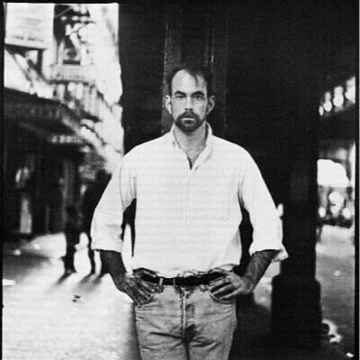 A man with a beard and glasses standing on a city street with hands on hips