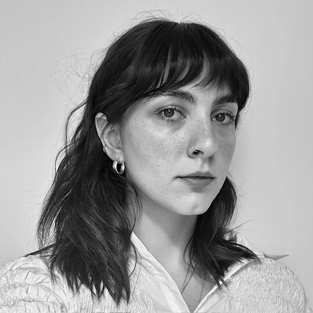 Black and white portrait of a woman with dark hair, nose ring, earrings, and a white textured shirt, looking at the camera.