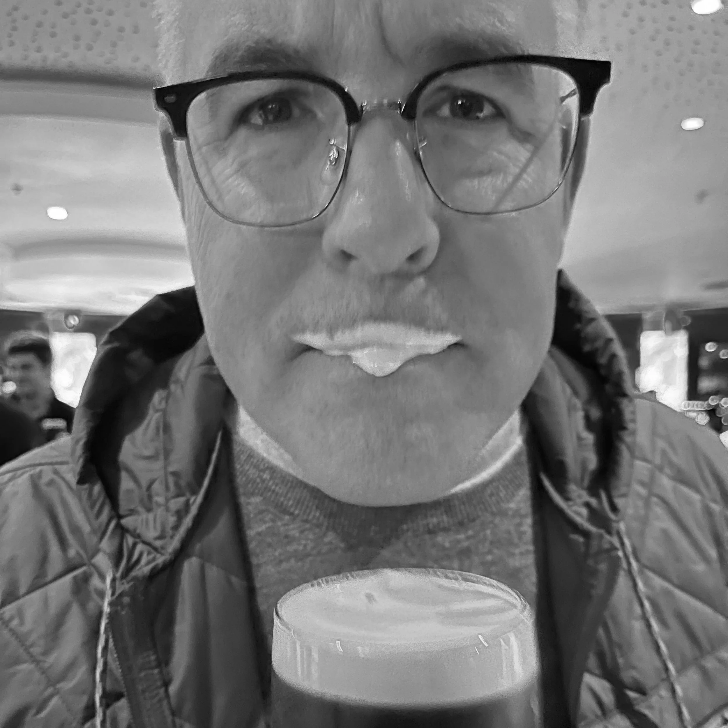 A close-up black and white photo of a man wearing glasses and a puffer jacket, holding a cup. The man is looking directly at the camera with a slight pursed lip.