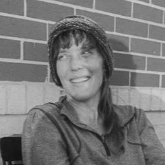 A woman with dark hair smiling, wearing a hat and a jacket, sitting against a brick wall.