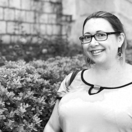 A woman with glasses smiling outdoors, wearing a light-colored top with dark trim, in front of plants and a stone wall.