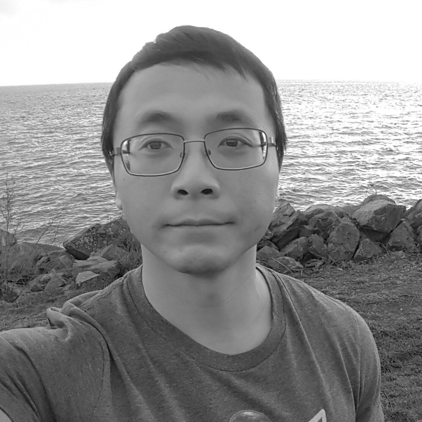 A black and white photo of a young person with glasses, short dark hair, and a neutral expression, standing outdoors near a rocky shoreline with water in the background.