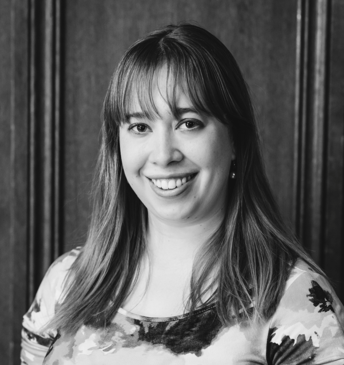 Black and white portrait of a woman with shoulder-length hair, smiling, wearing earrings and a patterned top, standing in front of a wooden background.