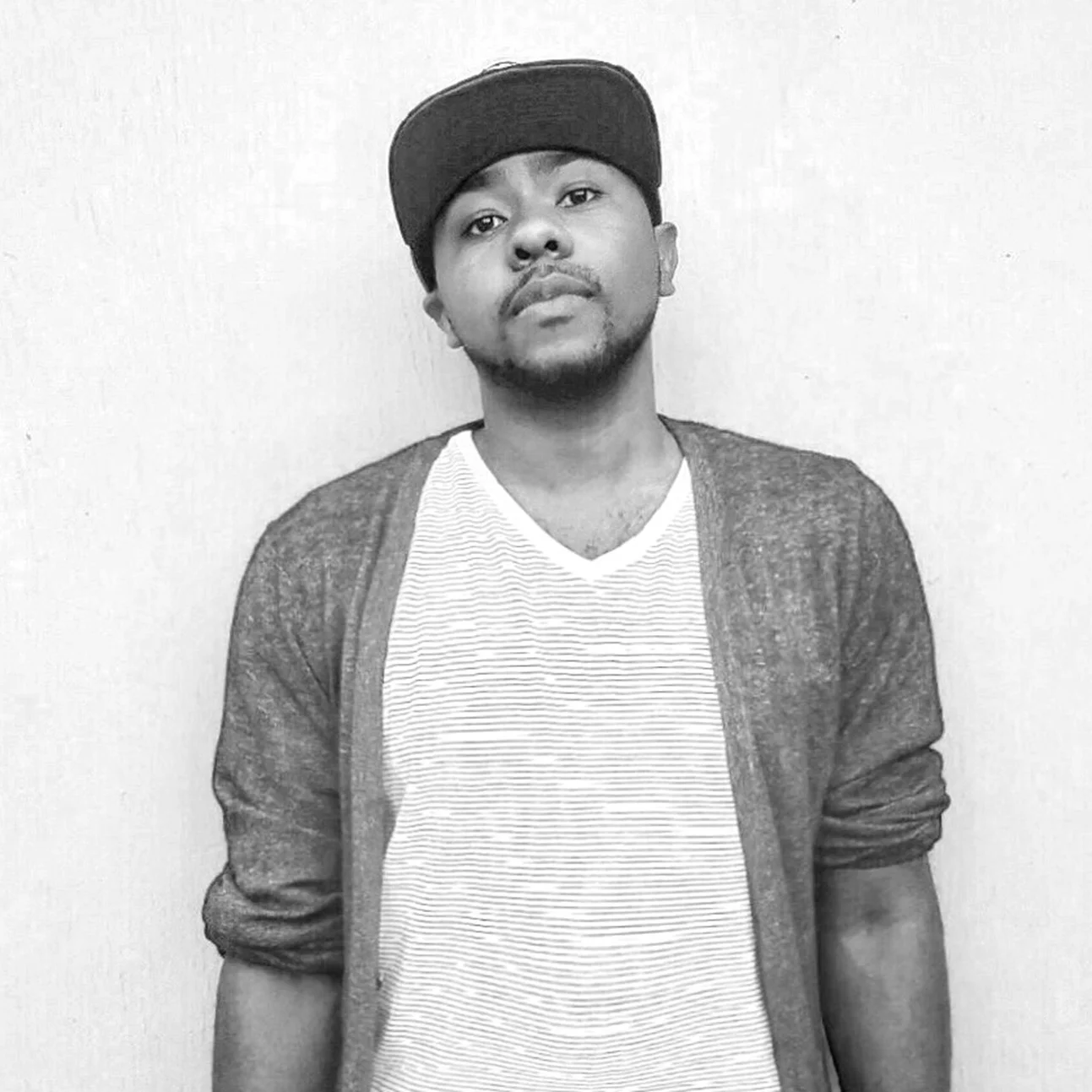 A young man in a baseball cap, striped t-shirt, and open cardigan posing against a plain background.