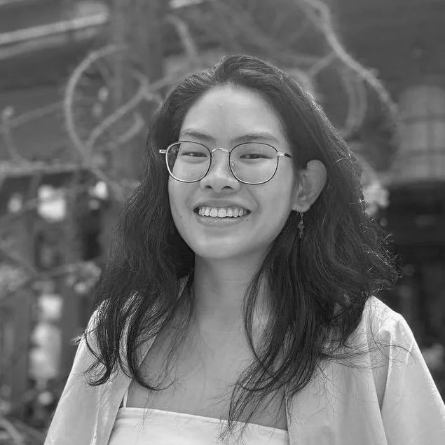 A young woman with glasses and wavy hair smiling outdoors in black and white.
