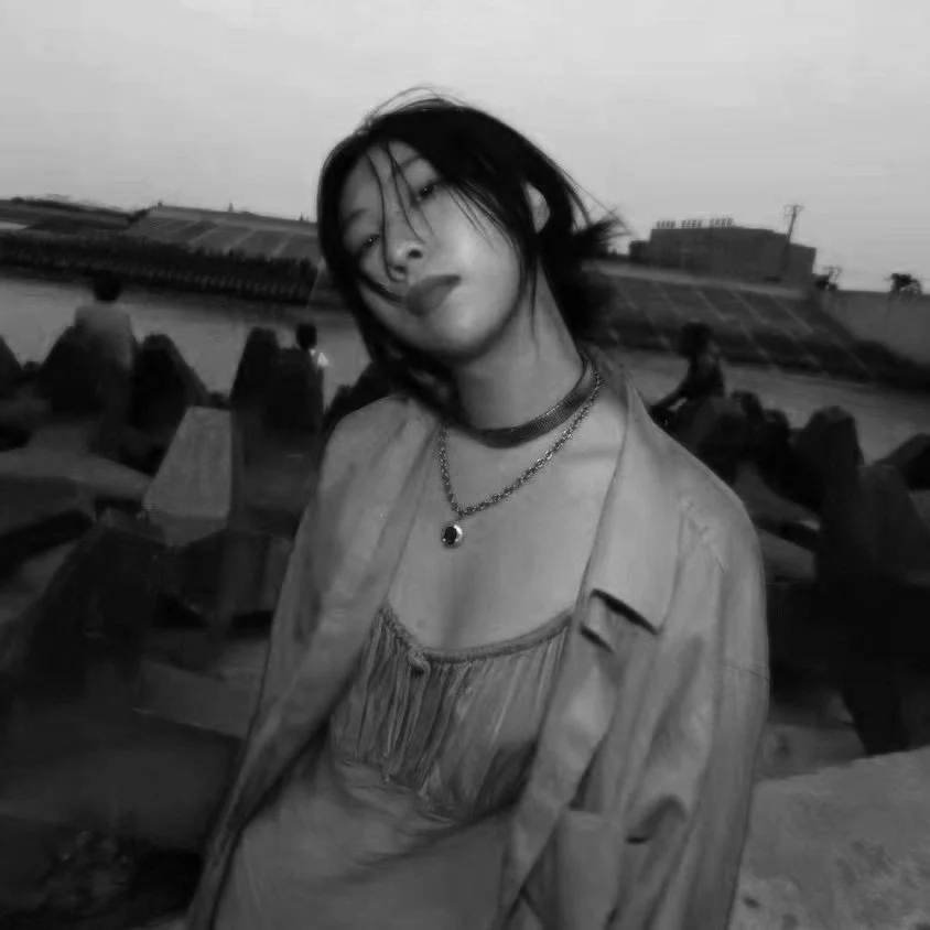 A young woman with short dark hair and layered clothing poses outdoors at sunset, with a harbor and boats in the background.