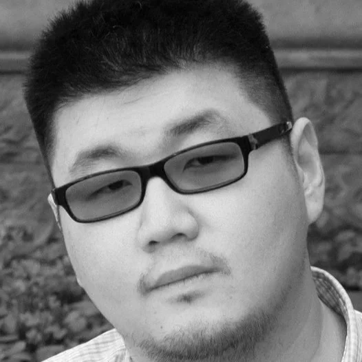 A young man with short, dark hair and glasses looking at the camera in black and white.