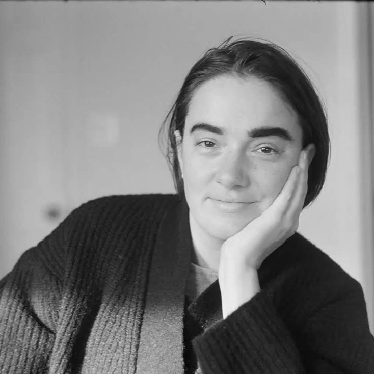Black and white photo of a young woman with short dark hair, resting her face on her hand, looking at the camera with a slight smile, wearing a dark sweater.
