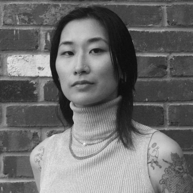 Black and white photo of a young woman with shoulder-length hair, wearing a turtleneck sweater and layered necklaces, standing in front of a brick wall.