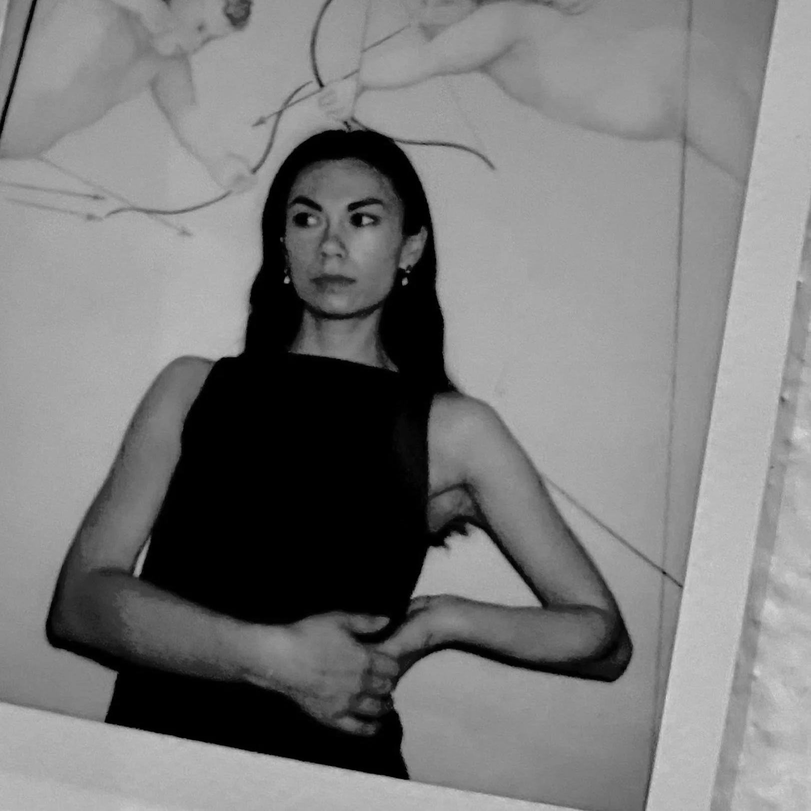 Black and white photo of a woman wearing a sleeveless dress, standing with her hands clasped in front of her, looking to her left with a serious expression. Background includes a wall with a drawing or painting of a flower or plant.