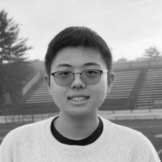 Black and white photo of a young man with short hair and glasses smiling outdoors, with bleachers and trees in the background.