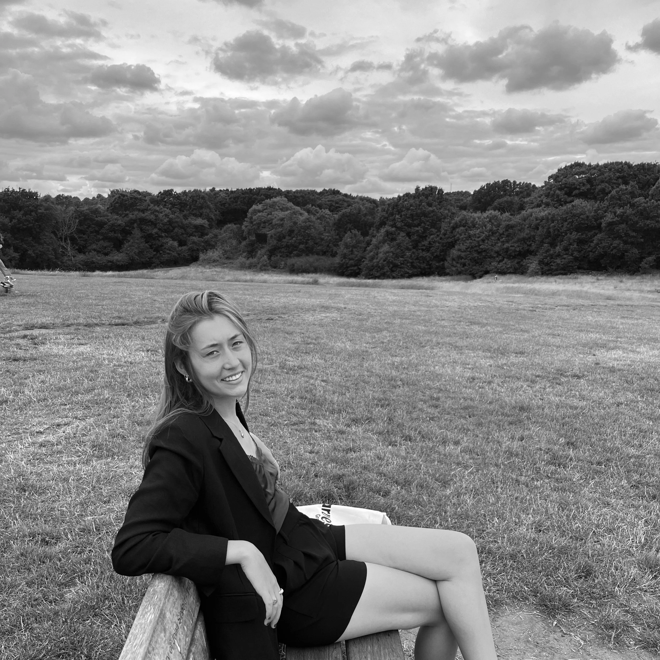 A young woman in business attire sitting on a park bench in a grassy open field with trees and clouds in the background, smiling at the camera.