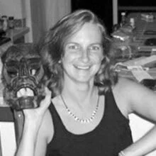 A woman with shoulder-length hair, smiling, holding a tiger head mask or sculpture, in a room with various objects and papers in the background.