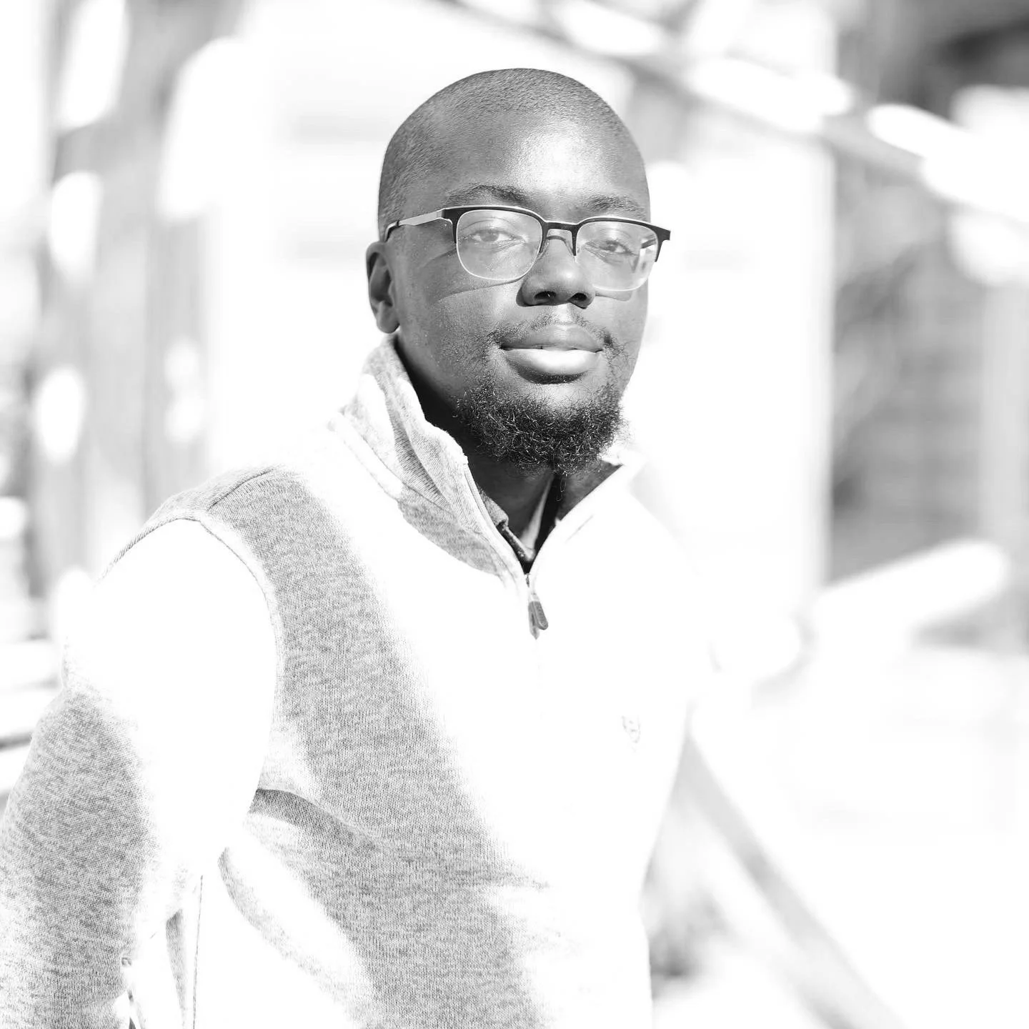 Black and white portrait of a man with glasses, a short beard, and a shaved head, wearing a zip-up sweater, outdoors.