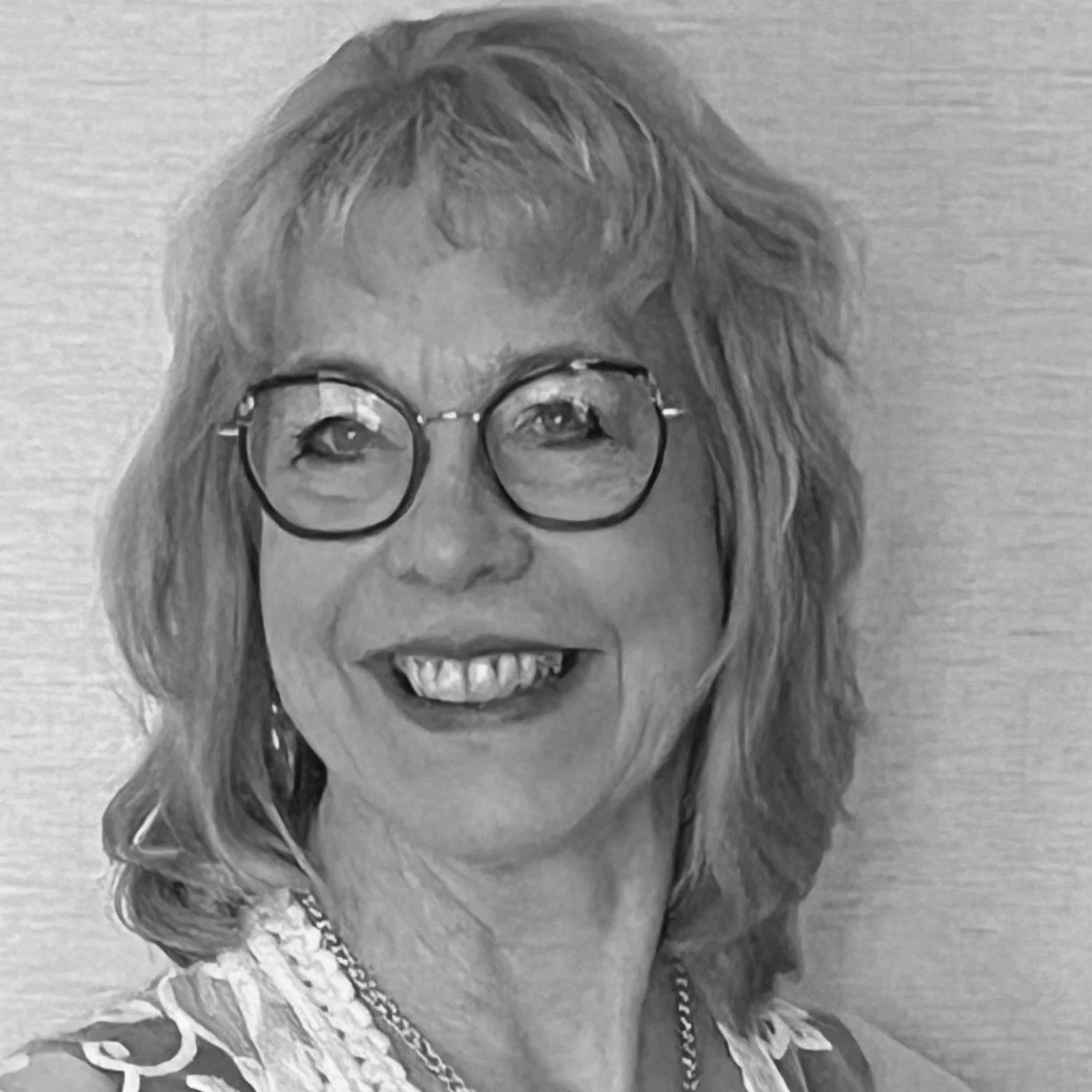 Black and white photo of a woman with shoulder-length hair, wearing glasses, smiling, and dressed in a patterned top with a chain necklace.