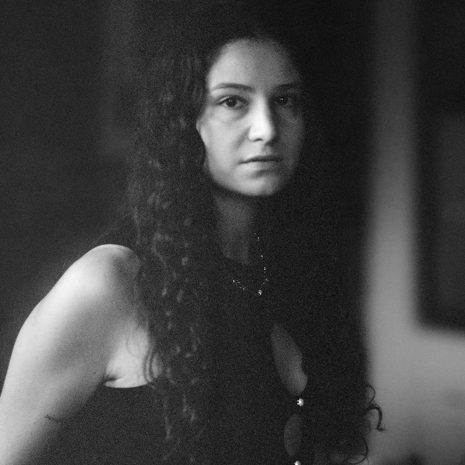A black-and-white portrait of a woman with long, curly hair looking directly at the camera with a serious expression.