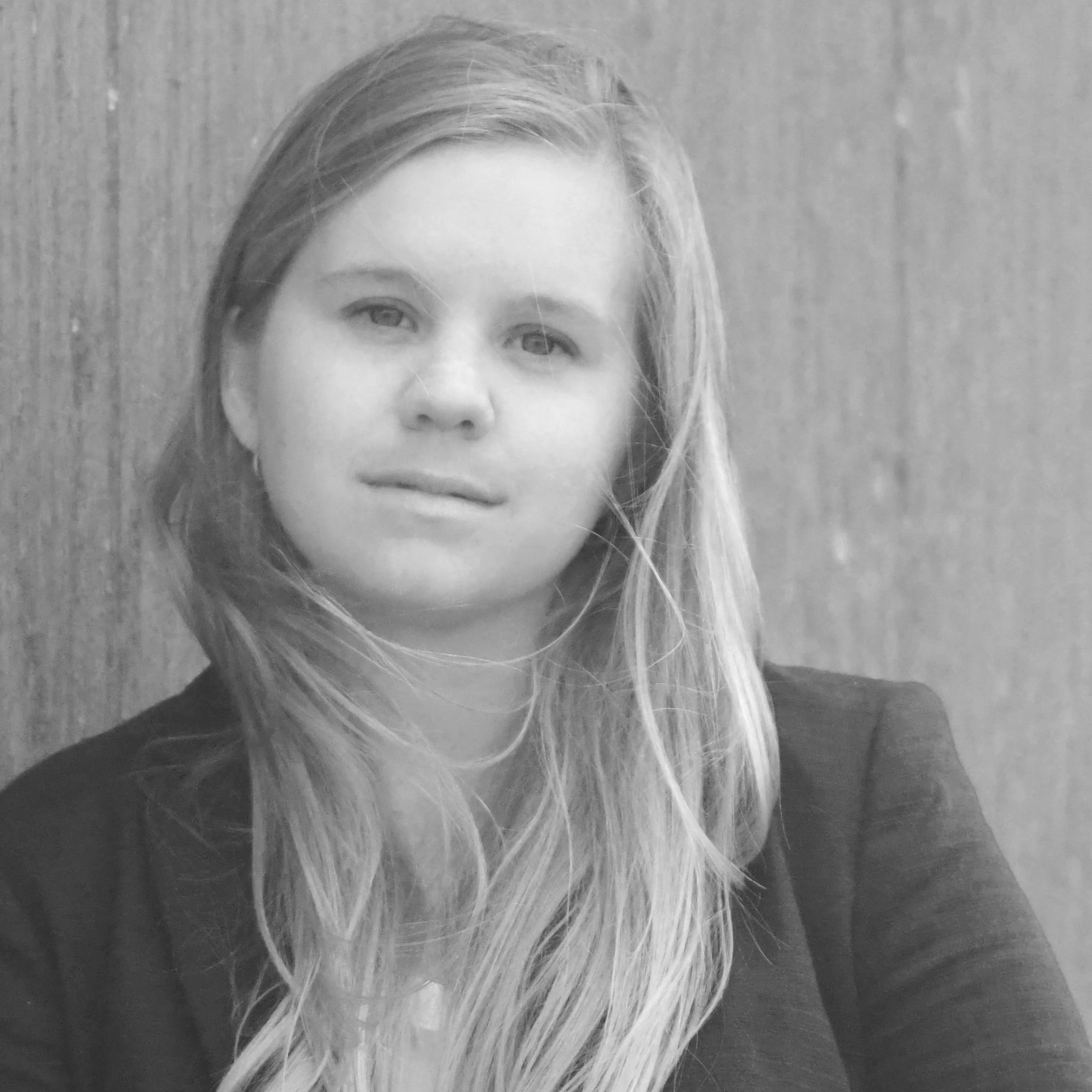 Black and white portrait of a woman with long hair, wearing a blazer, against a wooden background.