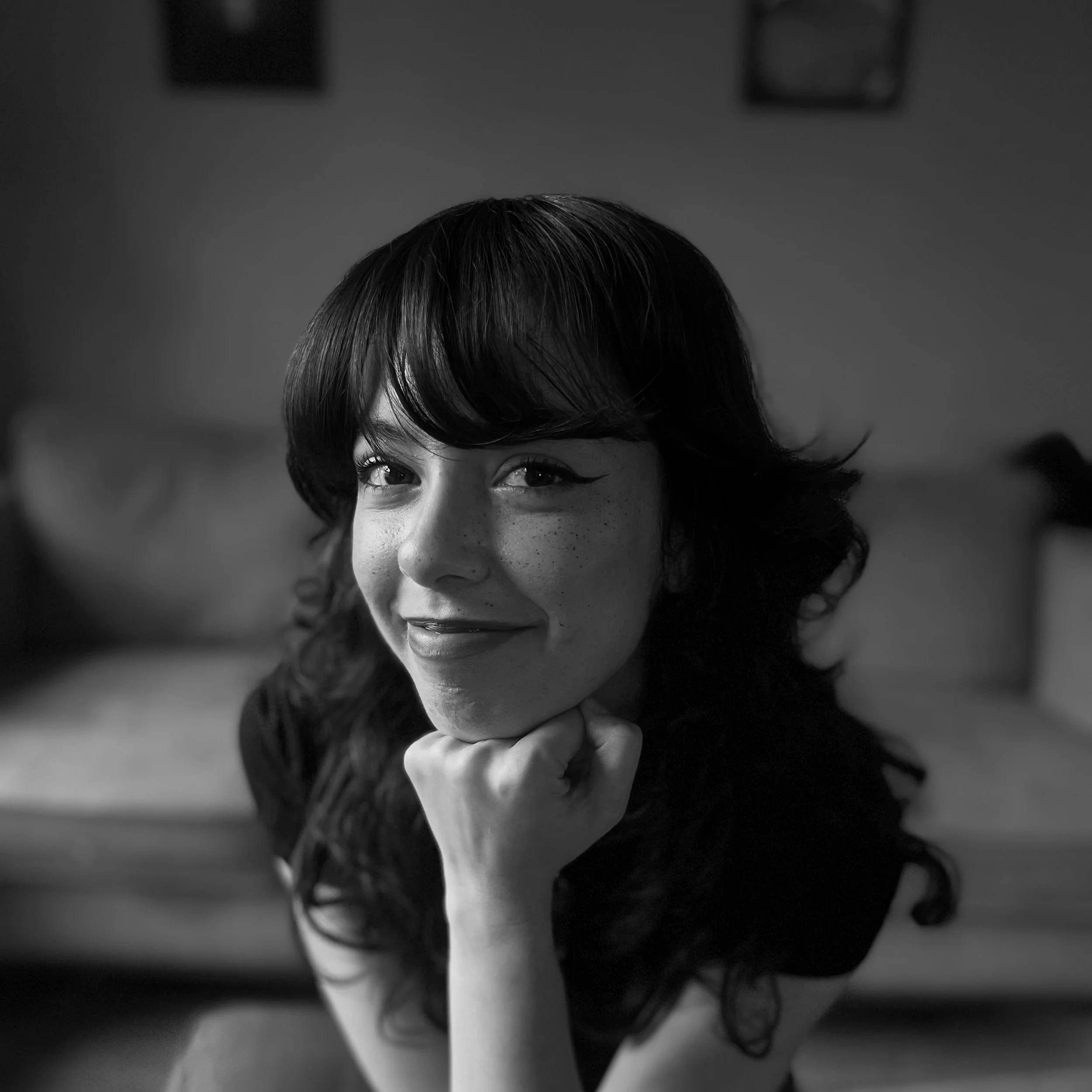 Black and white portrait of a woman with wavy hair, resting her chin on her hand, smiling thoughtfully indoors.