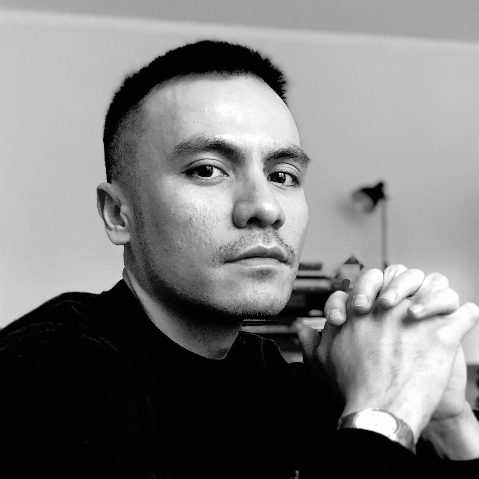Black and white portrait of a man with short dark hair, sitting at a desk with hands clasped, looking directly at the camera with a serious expression, in an indoor setting with a lamp and books in the background.