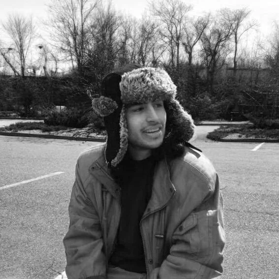 A young man in a winter coat and a furry hat with earflaps, smiling and sitting outdoors in a parking lot with leafless trees in the background.