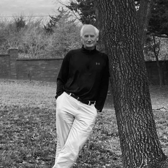 An older man with white hair, wearing a black long-sleeve shirt and white pants, leaning against a tree in a yard with a brick fence in the background.