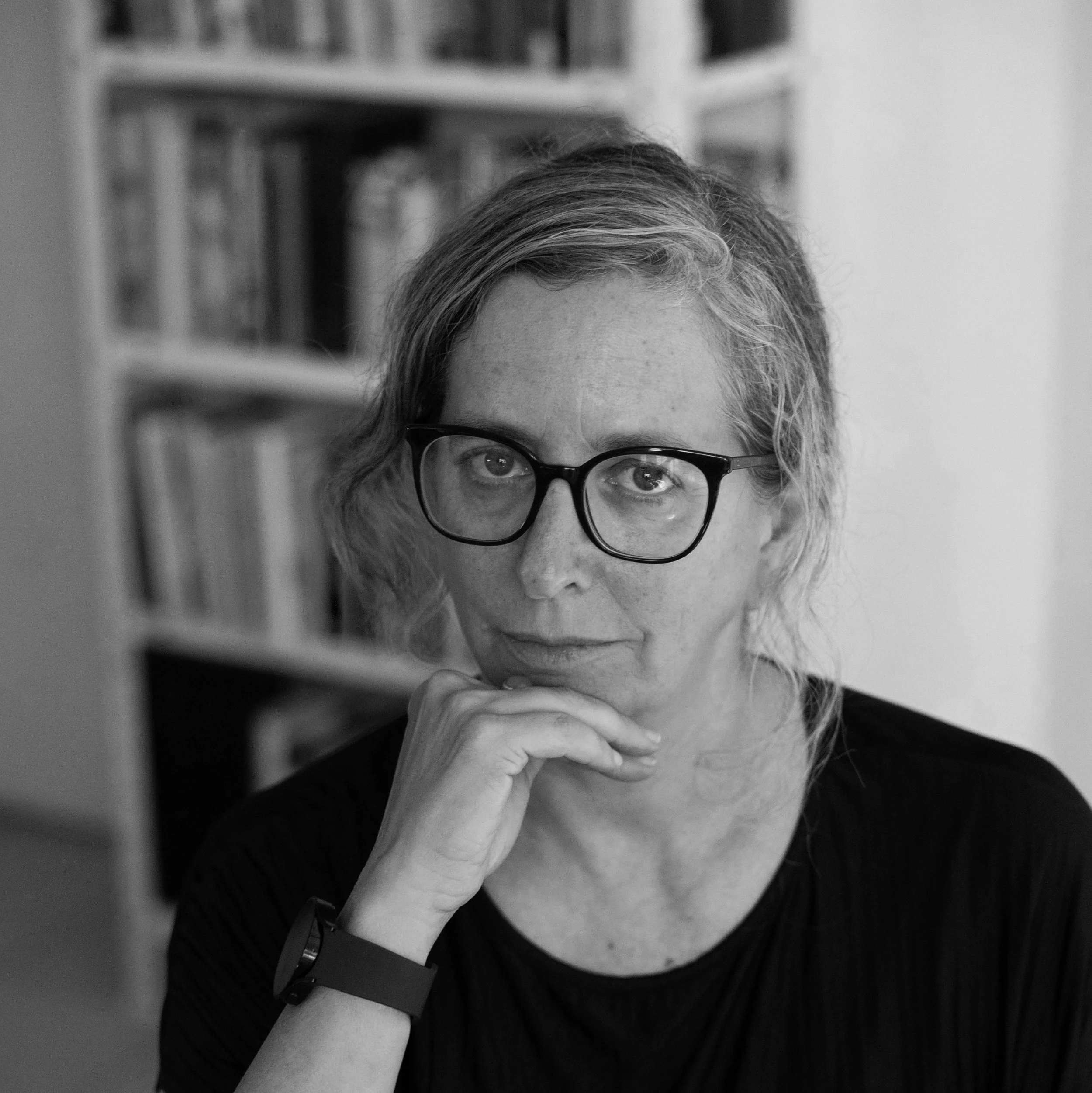 A woman with curly hair, glasses, and freckles, resting her chin on her hand, in front of bookshelves.