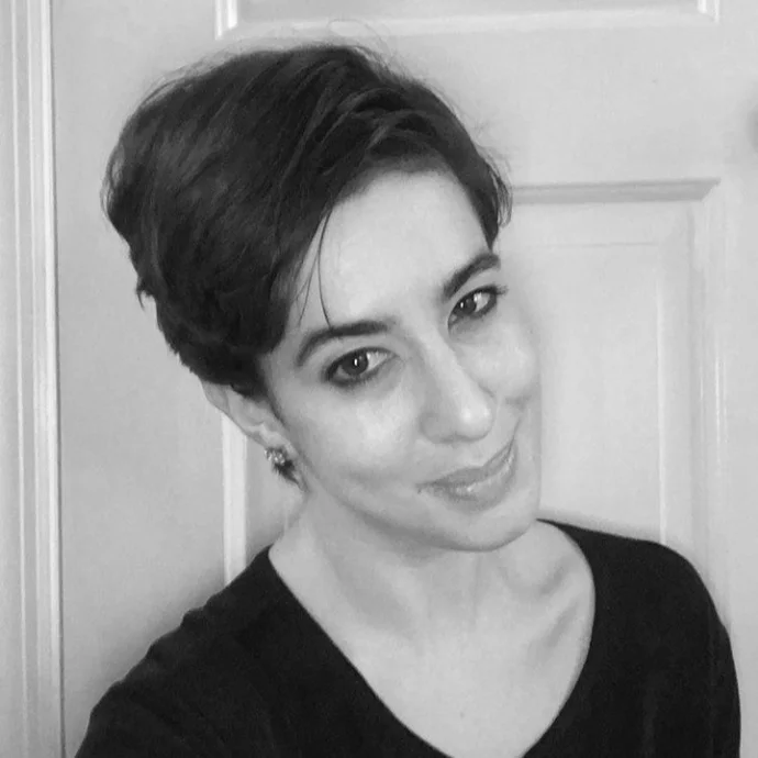 Black and white photo of a woman with short dark hair, smiling slightly, wearing a dark top, standing in front of a door.