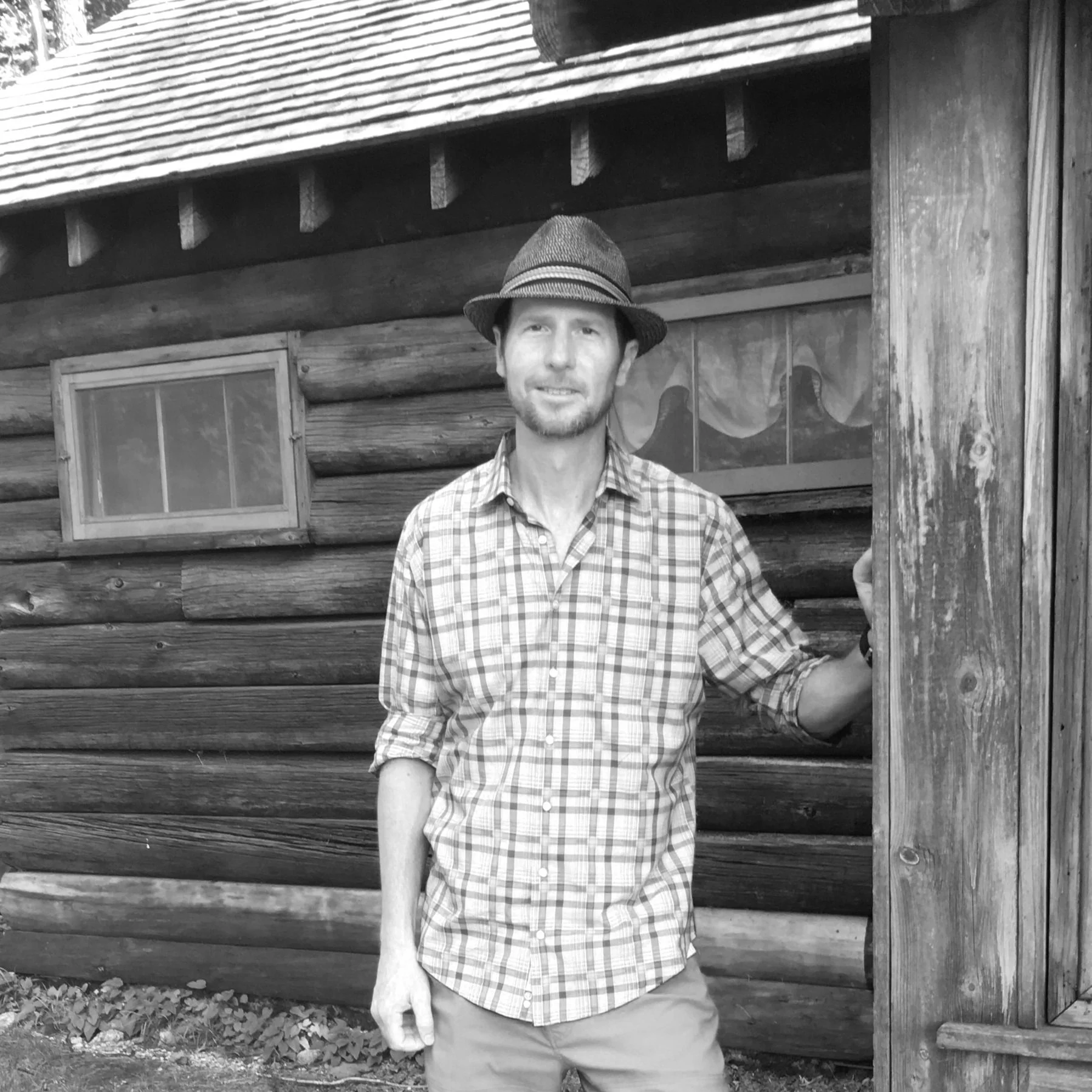 A man in a plaid shirt and hat standing in front of a log cabin, holding the door frame with one hand and smiling.