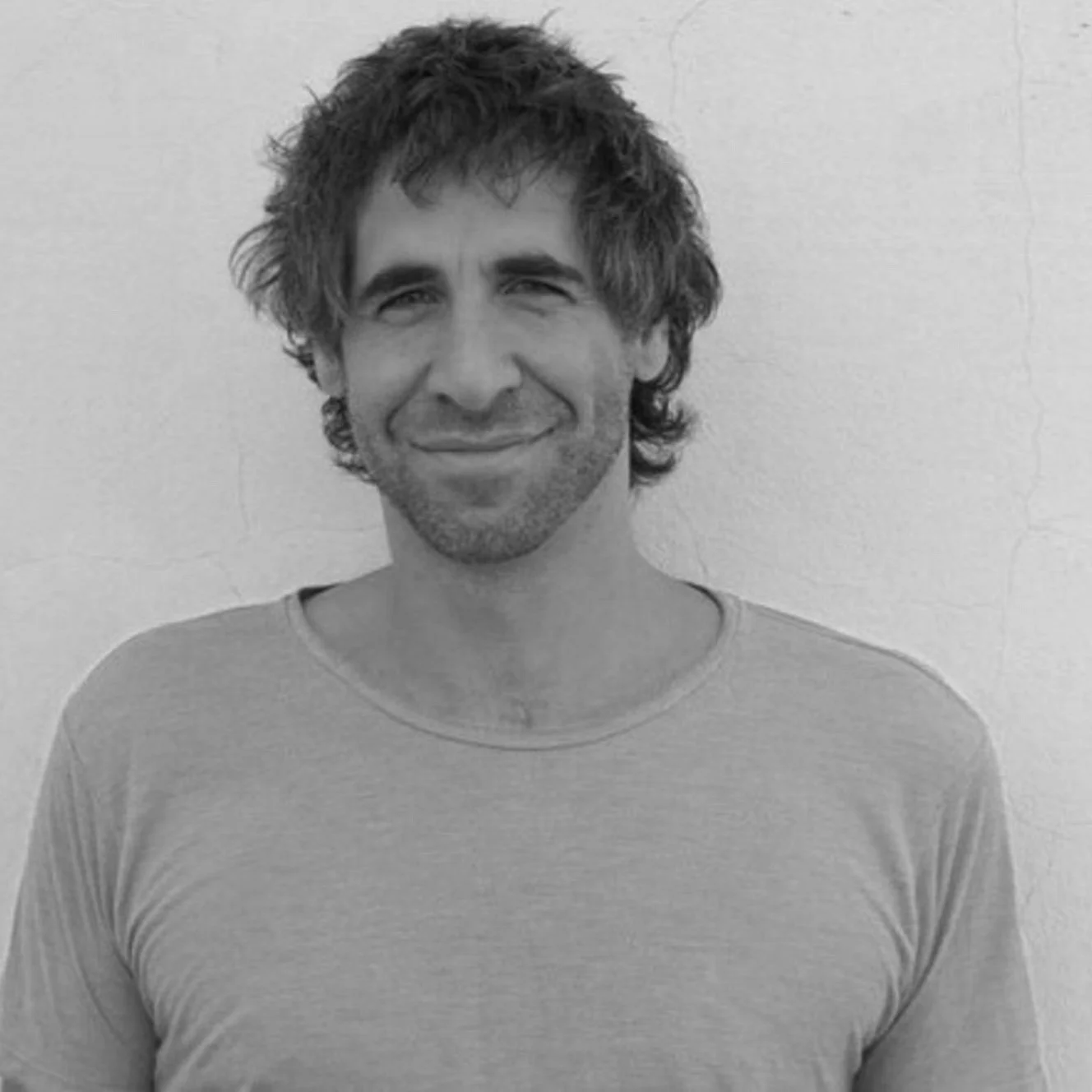 A black and white photo of a man with curly hair and a beard, smiling and wearing a plain t-shirt, standing against a plain wall.