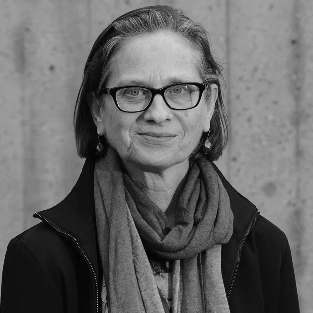 Black and white portrait of a woman with glasses, earrings, a scarf, and a zip-up jacket, standing against a plain wall.