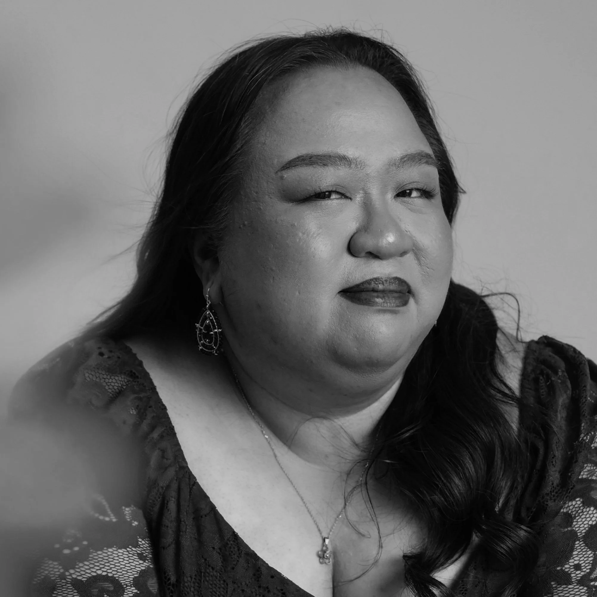 Black and white portrait of a woman with long dark hair, wearing earrings, a necklace, and a lace top, looking slightly to the side.