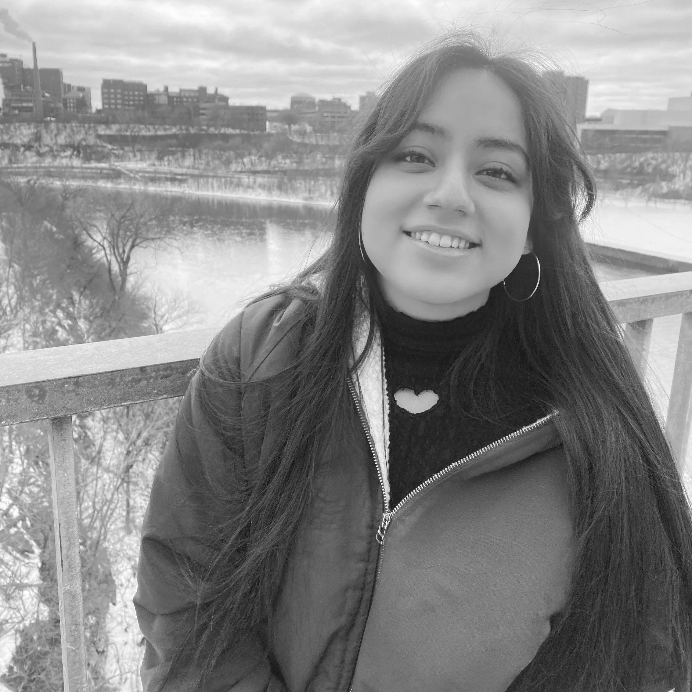 A woman with long dark hair wearing hoop earrings, a heart-shaped necklace, a black turtleneck, and a jacket, smiling outdoors with a city skyline and a river in the background.